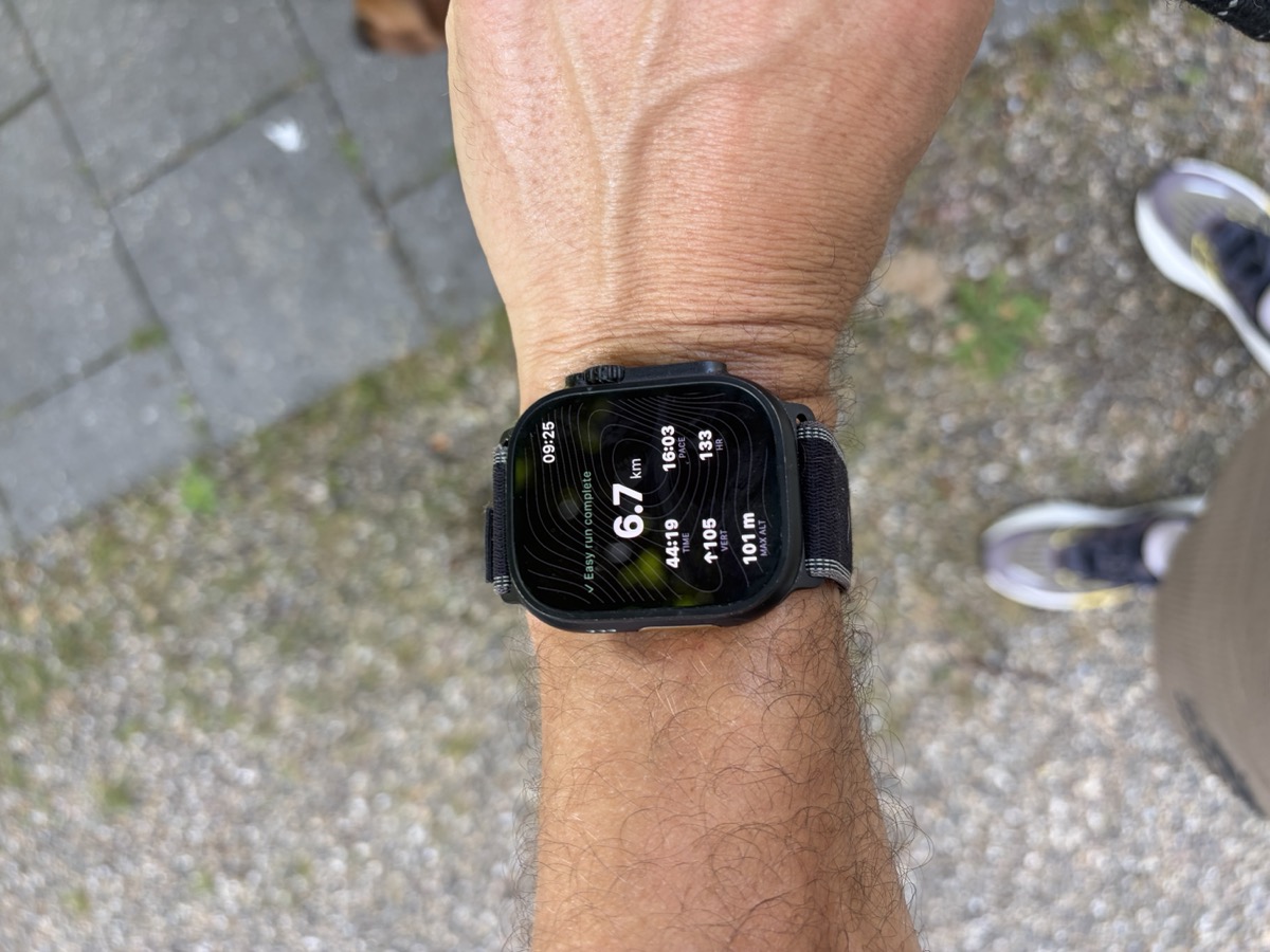 An Apple Watch on the founder's wrist showing a completed easy run in Peak Days: 6.7 km, time, pace, vertical gain and heart rate over a topographic background.